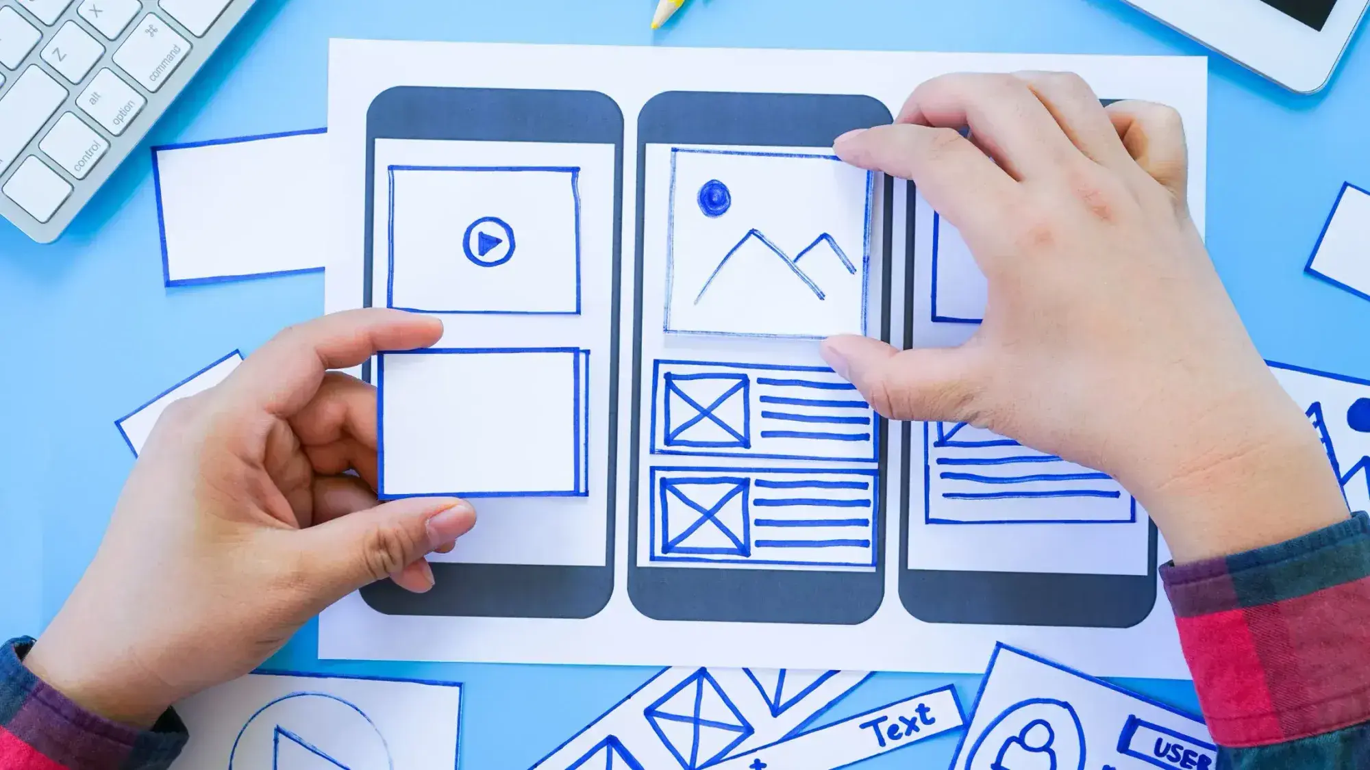 Hands arranging paper UI elements on a smartphone wireframe sketch. Includes icons, text boxes, and layout sections, conveying creativity and design planning.